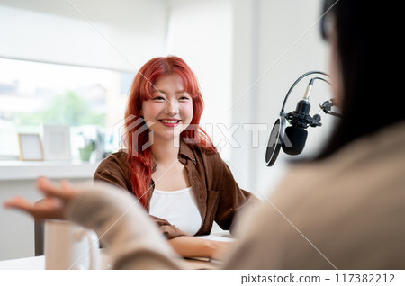 A friendly Asian female podcaster is enjoying a conversation as she interviews a guest in her studio 117382212