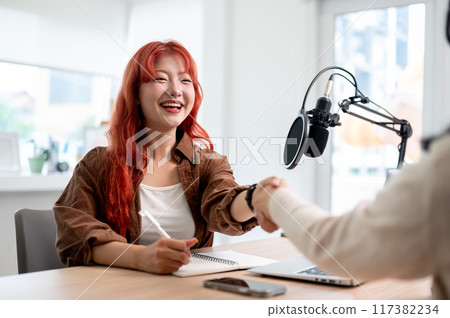 A friendly Asian female podcaster is shaking hands with a guest, greeting them to the podcast show. 117382234