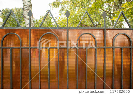 A fragment of a fence made of steel pipes and polycarbonate to protect private property, a summer day A fragment of a fence made of steel pipes and polycarbonate to protect private property, a summer day 117382690