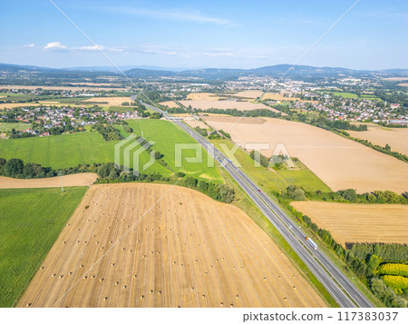 Aerial shot of golden hay bales scattered across fields adjacent to a busy highway, surrounded by lush green landscapes on a sunny day. 117383037