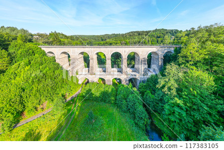 The Sychrov Railway Bridge stands majestically with graceful arches, surrounded by lush greenery. The bright sky enhances the tranquil setting. 117383105