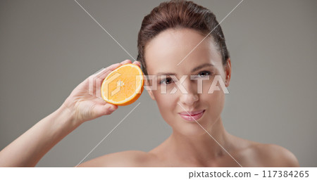 Beauty, health, cosmetics, anti-aging therapy and skin care concept - young beautiful brunette Caucasian woman holding half a orange, concept of freshness, vitamins and nutrients 117384265