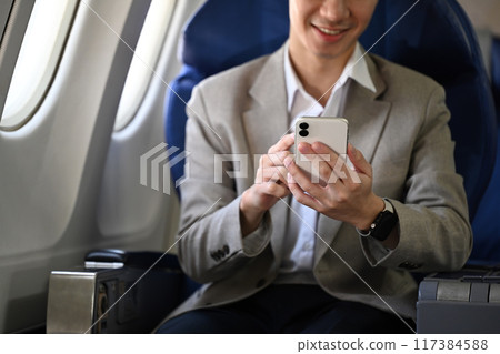Smiling young businessman sitting with smartphone on the aircraft seat near the window during the flight 117384588