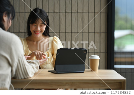 Cheerful young female colleagues sharing something on mobile phone while sitting in the modern cafe 117384822
