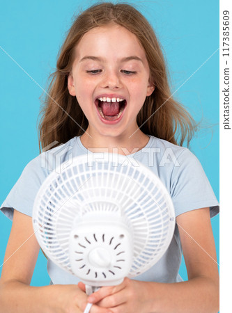 Child girl enjoying cool air. Child and fan on blue background. Room temperature regulation 117385609