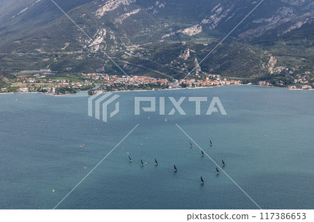 Fleet of sailboats racing on sparkling Lake Garda, with lush Italian countryside and mountains 117386653