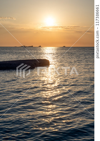 Vertical photo of Cala Conta beach sunset, low sun casting reflections on dark, textured sea surface 117386661
