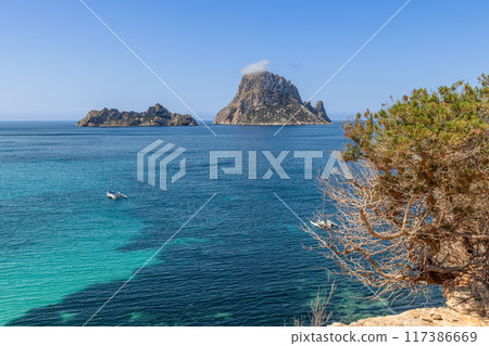 Tranquil scene of Es Vedra, Ibiza, with catamaran anchored in turquoise waters, cliffs, and blue sky 117386669