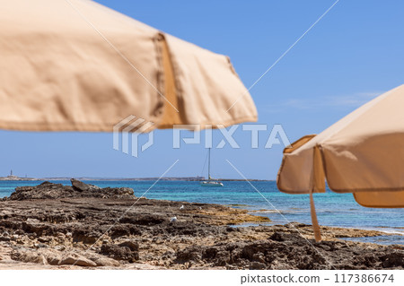 Beach umbrellas on rocky Ibiza shore with clear emerald sea, yacht sailing distant Formentera island Beach umbrellas on rocky Ibiza shore with clear emerald sea, yacht sailing distant Formentera island 117386674