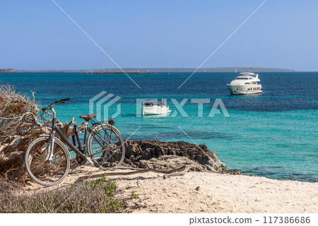 Bicycle leans against driftwood on Ibiza coast, with luxury yachts sailing in clear blue waters Bicycle leans against driftwood on Ibiza coast, with luxury yachts sailing in clear blue waters 117386686