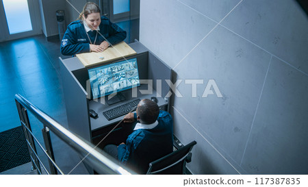 African American male security guard sits at the workplace with PC, talks to female colleague African American male security guard sits at the workplace with PC, talks to female colleague 117387835