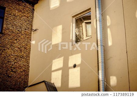 Window in yellow wall with reflections of other windows on the wall as well as a water pipe and a brick wall as a corner building 117389119