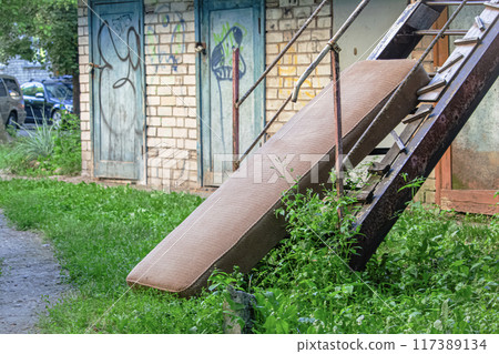 Old brown textured mattress leaning on metal stairs left outside near brick wall shed with wooden doors and green grass around Old brown textured mattress leaning on metal stairs left outside near brick wall shed with wooden doors and green grass around 117389134