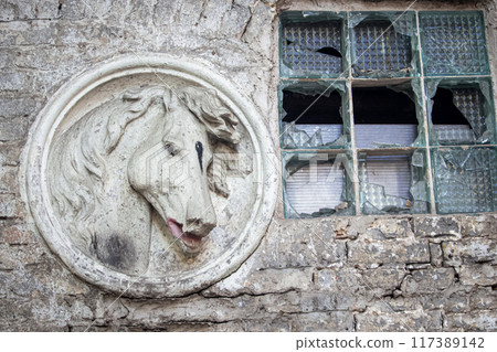 White horse's head sculpture near a broken thick glass window in weathered brick wall 117389142