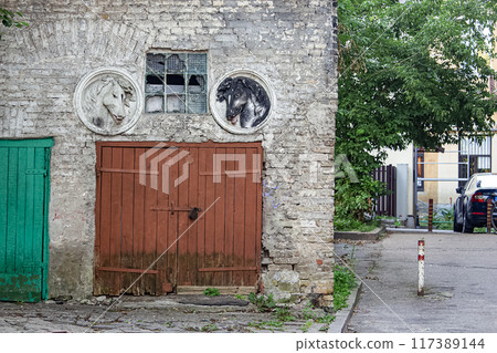 Old and worn brown wooden garage door of a former horse stable with sculptures of horses' heads over it and a thick glass window Old and worn brown wooden garage door of a former horse stable with sculptures of horses' heads over it and a thick glass window 117389144