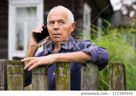 Elderly man standing beside his house and talking on phone Elderly man standing beside his house and talking on phone 117390443