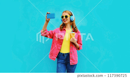happy smiling woman taking selfie with phone listening to music in headphones in pink jacket on blue 117390725