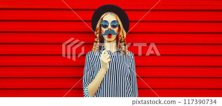 Portrait of funny cheerful young woman having fun showing mustache on stick on red background Portrait of funny cheerful young woman having fun showing mustache on stick on red background 117390734