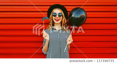 Portrait of funny cheerful woman having fun holding black balloon, showing mustache on stick on red 117390735