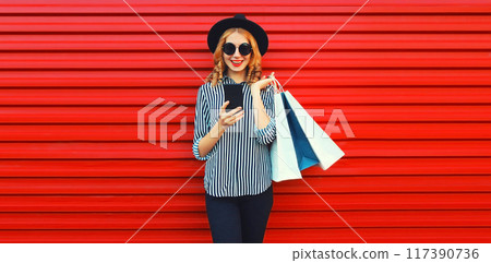 Portrait of happy smiling woman with shopping bags and phone black round hat on red background Portrait of happy smiling woman with shopping bags and phone black round hat on red background 117390736