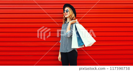 Portrait of smiling young woman with shopping bags in striped shirt, black hat on red background Portrait of smiling young woman with shopping bags in striped shirt, black hat on red background 117390738