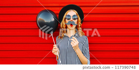 Portrait of funny cheerful woman having fun holding black balloon, showing mustache on stick on red 117390748