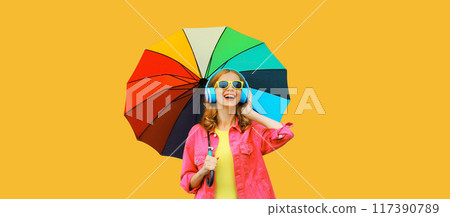 Happy cheerful young woman with colorful umbrella listening to music in headphones on yellow 117390789