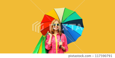 stylish happy smiling young woman holding colorful umbrella with shopping bags on yellow background 117390791