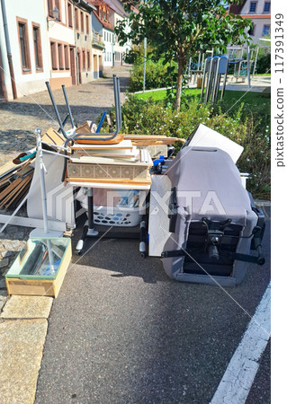 Discarded items including overturned office chair, wooden boards, home furnishings cluttering sidewalk, reflecting urban waste management issues, necessity for responsible disposal. Vertical picture. 117391349