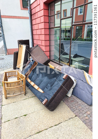 A pile of discarded furniture including a sofa, chair, and various cushions on a city sidewalk, highlighting urban waste and the need for proper disposal or recycling. Vertical picture. A pile of discarded furniture including a sofa, chair, and various cushions on a city sidewalk, highlighting urban waste and the need for proper disposal or recycling. Vertical picture. 117391361