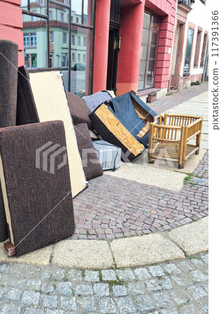 A pile of discarded furniture including a sofa, chair, and various cushions on a city sidewalk, highlighting urban waste and the need for proper disposal or recycling. Vertical picture. A pile of discarded furniture including a sofa, chair, and various cushions on a city sidewalk, highlighting urban waste and the need for proper disposal or recycling. Vertical picture. 117391366