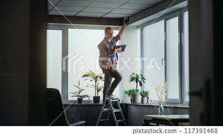 Male technician stands on ladder, installs CCTV camera in office room using tablet Male technician stands on ladder, installs CCTV camera in office room using tablet 117391468