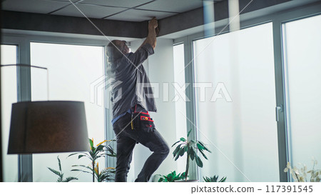Male technician stands on ladder, installs CCTV camera in office room using tablet 117391545