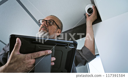 Male installer adjusts the angle of CCTV camera in office, uses tablet computer 117391584