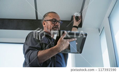 Male mature installer sets up CCTV camera in office room using tablet computer Male mature installer sets up CCTV camera in office room using tablet computer 117391590