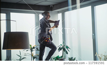 Male technician stands on ladder, installs CCTV camera in office room using tablet 117391591