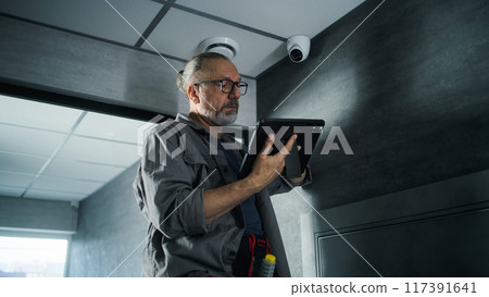 Male installer adjusts the angle of CCTV camera in office, uses tablet computer 117391641
