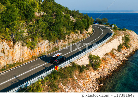 A black car with a paddleboard drive along a narrow coast with clear water and a road under a clear blue sky. 117391642