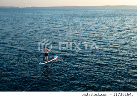Man in orange vest navigating on the paddleboard in the sea at sunset. 117391643