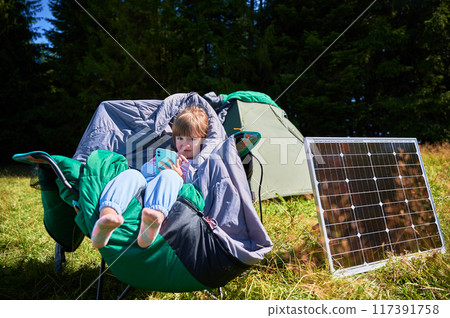 Young child plays with smartphone while charging with photovoltaic solar panel near tourist tent in summer. Integration of renewable energy in outdoor camping activities. Young child plays with smartphone while charging with photovoltaic solar panel near tourist tent in summer. Integration of renewable energy in outdoor camping activities. 117391758