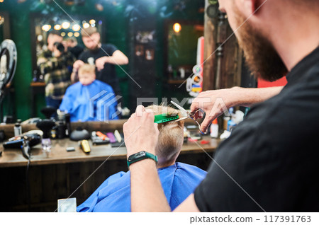 Behind shoulder view of male barber cutting little boy's hair. Young hairdresser using comb and scissors to cut straight. Professional hairdresser and cute client in front of big mirror. 117391763