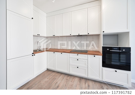 Kitchen with laminate floor, white walls, doorway and modern kitchen set with electric stove, counter, sink and oven. Apartment kitchen room with elegant minimalist design in white colors. Kitchen with laminate floor, white walls, doorway and modern kitchen set with electric stove, counter, sink and oven. Apartment kitchen room with elegant minimalist design in white colors. 117391782