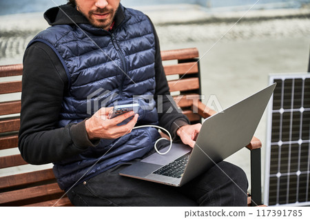 Cropped view of man sits on wooden bench, working on laptop and smartphone, both connected to solar panel. Urban setting and casual attire highlight modern, sustainable, mobile workspace. 117391785