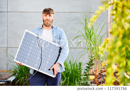 Portrait of bearded man holds solar panel, standing in modern setting with plants and greenery. Guy in light blue shirt and white t-shirt looks happy and relaxed. 117391786