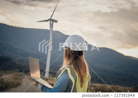 A skilled professional woman oversees a complex wind turbine operation on her laptop, surrounded by nature, showcasing the fusion of advanced technology and environmental awareness 117391949