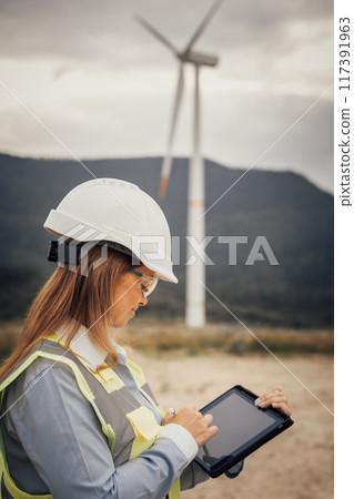 A skilled female engineer uses a modern tablet at a vast wind farm, highlighting the blend of advanced technology and innovative engineering in a sustainable, ecofriendly environment 117391963