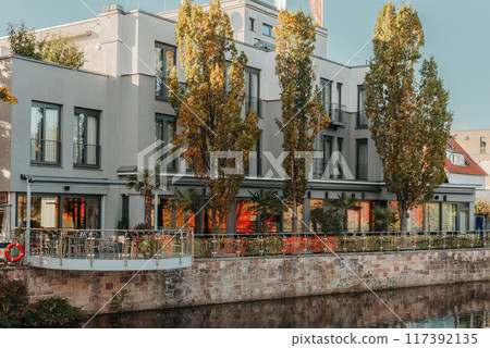 River On Sunset. Townhouses Buildings And Multi-floor Home At Waterfront. Sunset On River In City. Suburb Houses And Residential Building Near River In Europe. House In Nature On Shore On Sunset. 117392135