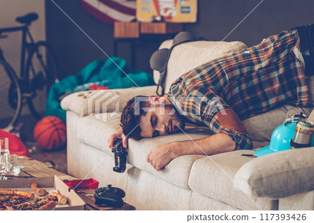 Young handsome man passed out on sofa with joystick in his hand in messy room after party 117393426