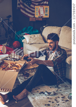 Excited young man holding joystick and keeping mouth open while sitting on the floor in messy room after party Excited young man holding joystick and keeping mouth open while sitting on the floor in messy room after party 117393437