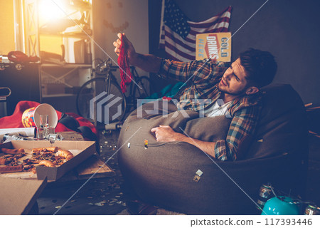 Handsome young man holding red lace panties and looking at it while lying on bean bag in messy room after party Handsome young man holding red lace panties and looking at it while lying on bean bag in messy room after party 117393446